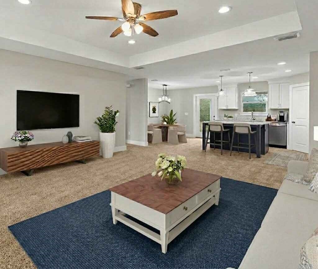 Modern open-concept living room with wood TV console, ceiling fan, blue area rug, white coffee table, and kitchen visible in background